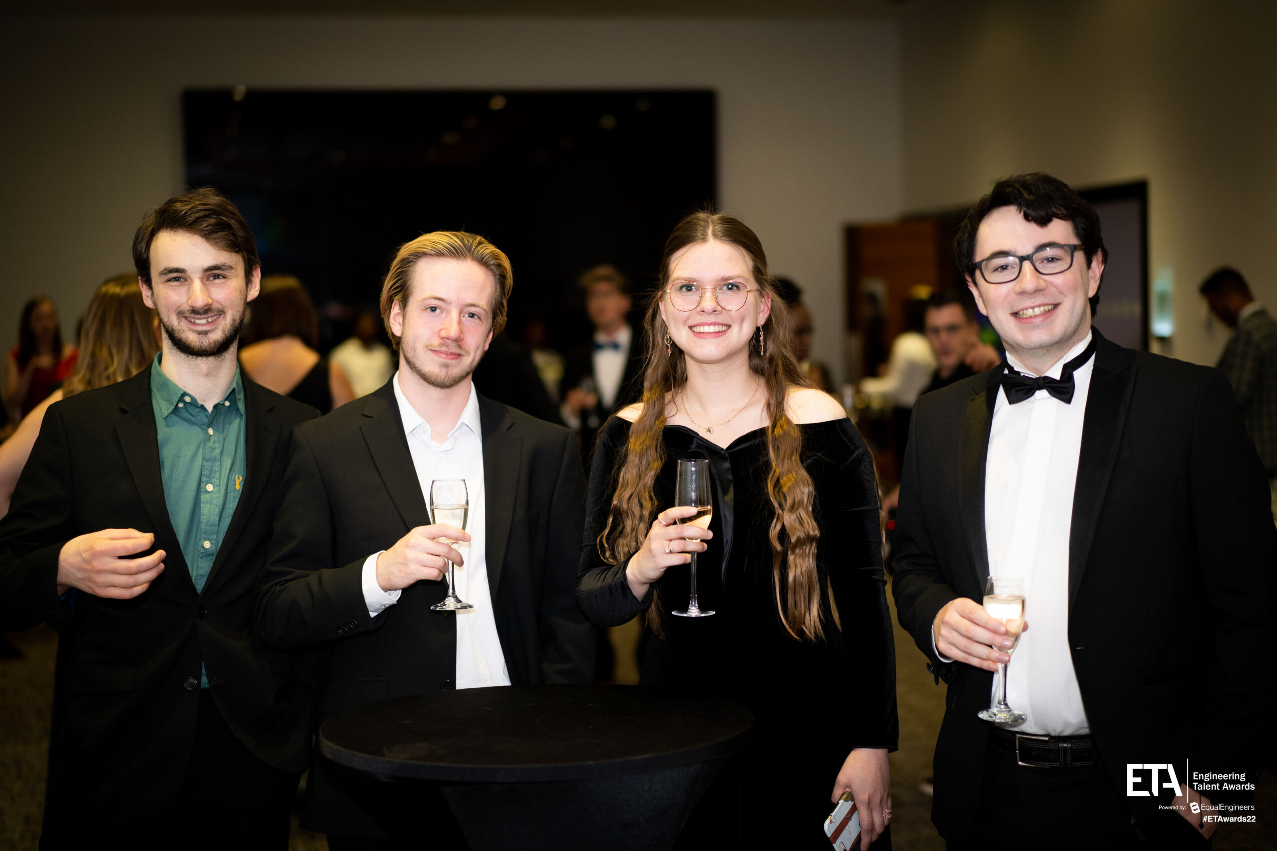 A group of 4 men and women in dinner dress hold drinks and face camera