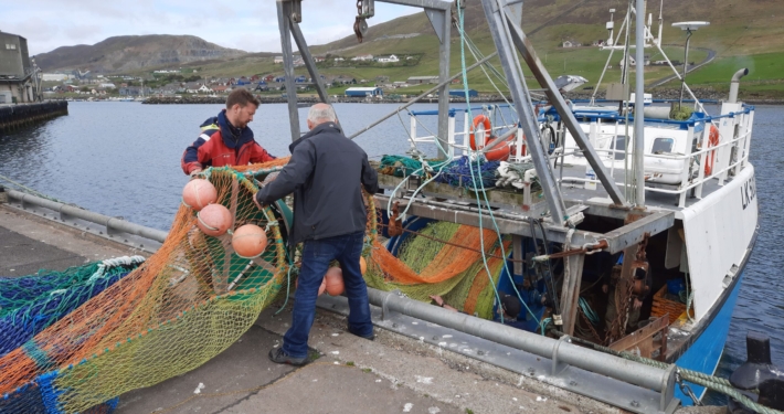 two white men stand at a dock load a fishing net into a medium-sized trawling vessel