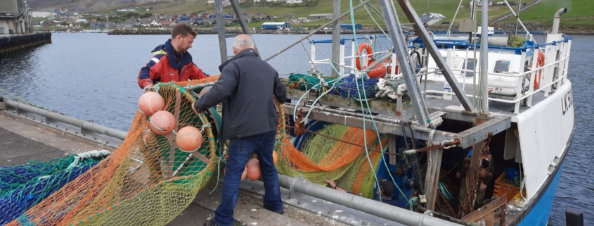 two white men stand at a dock load a fishing net into a medium-sized trawling vessel