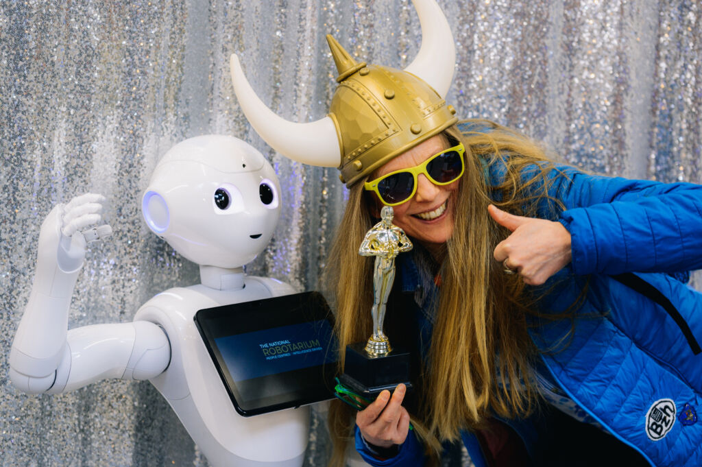 A blonde woman wearing a Viking hat and sunglasses stands smiling beside a humanoid robot