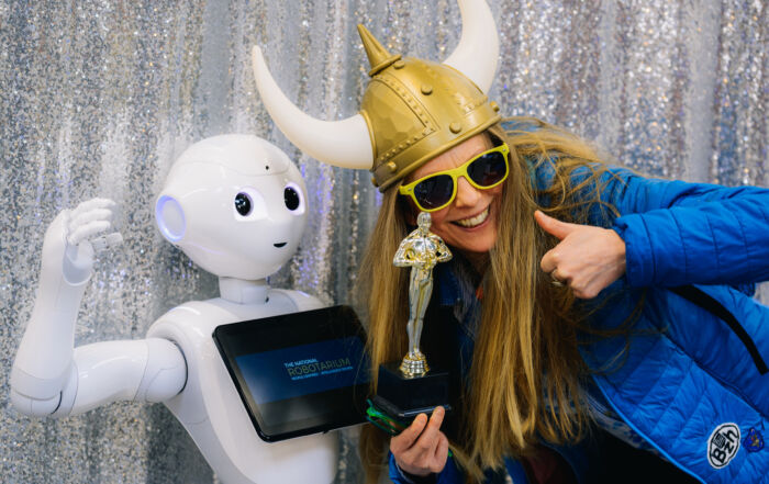 A blonde woman wearing a Viking hat and sunglasses stands smiling beside a humanoid robot