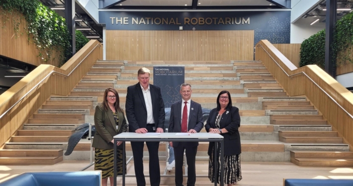 The image shows two women and two men standing at a table in front of a staircase. The background prominently displays the text "The National Robotarium." The individuals are posing for a formal photograph, involving a memorandum of understanding (MOU). The setting is modern with natural light, wooden accents, and greenery, indicative of a well-designed interior space.