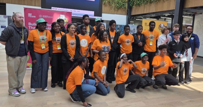 A large group of students and mentors pose together at a robotics competition event. Most of the students wear matching bright orange shirts with a logo, while others are in school uniforms. A banner in the background displays 'Inspire Award' and logos for FIRST Tech Challenge and the National Robotarium.