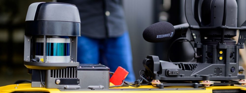 A person wearing a black shirt and blue jeans operates a handheld controller, focusing on a yellow robotic platform equipped with various sensors and devices. The robot, labeled "Boston Dynamics," features a lidar sensor, a mounted microphone, and a camera setup.