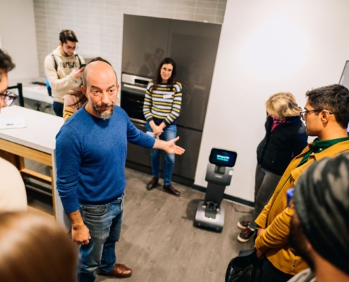 The image shows a group of people gathered in a room, where a man in a blue sweater is leading a demonstration or discussion. He is gesturing towards a small robot positioned on the floor.