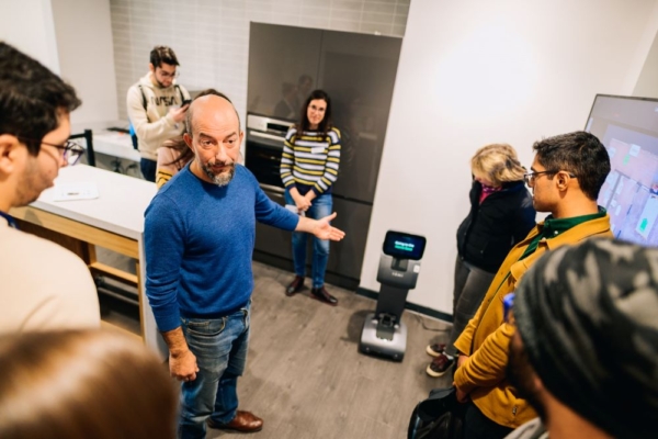 The image shows a group of people gathered in a room, where a man in a blue sweater is leading a demonstration or discussion. He is gesturing towards a small robot positioned on the floor.