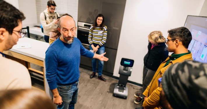 The image shows a group of people gathered in a room, where a man in a blue sweater is leading a demonstration or discussion. He is gesturing towards a small robot positioned on the floor.