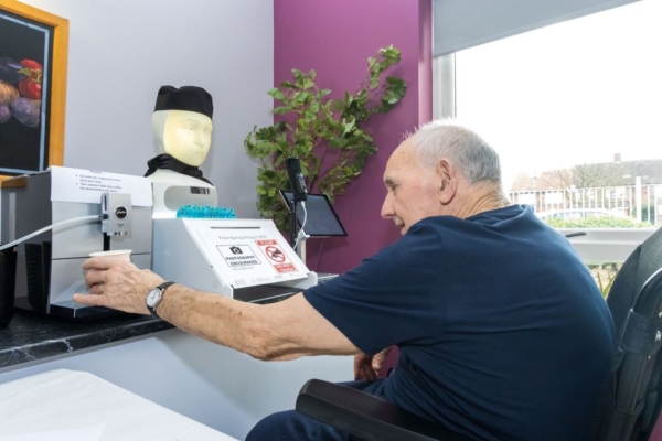 An elderly man is sitting in a wheelchair while interacting with a robotic coffee-making system labeled "Robo Barista," which has a humanoid head with a neutral expression, wearing a black cap and scarf. 