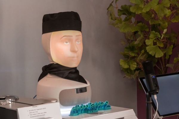 A close-up of the "Robo Barista 2025" robotic coffee-making system, featuring a humanoid head with a projected face, wearing a black cap and scarf.