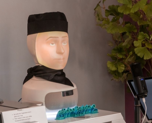 A close-up of the "Robo Barista 2025" robotic coffee-making system, featuring a humanoid head with a projected face, wearing a black cap and scarf.