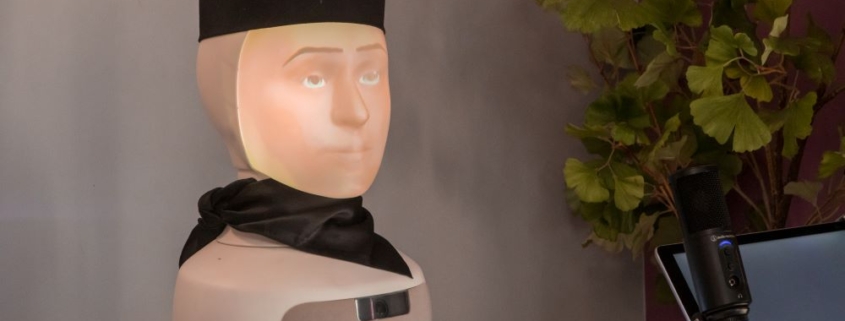 A close-up of the "Robo Barista 2025" robotic coffee-making system, featuring a humanoid head with a projected face, wearing a black cap and scarf.