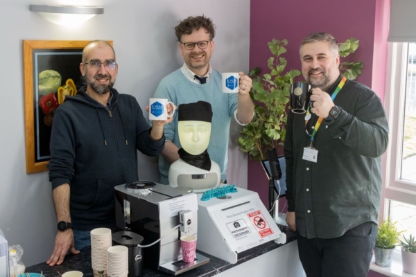 Three men stand together in front of a robotic coffee-making system called "Robo Barista," smiling and holding mugs.