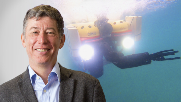 A middle-aged white man is visible from the shoulders up. He is wearing a blue shirt and brown jacket. In the background is an image of an underwater vessel with two large headlights. It is moving downwards in the sea.