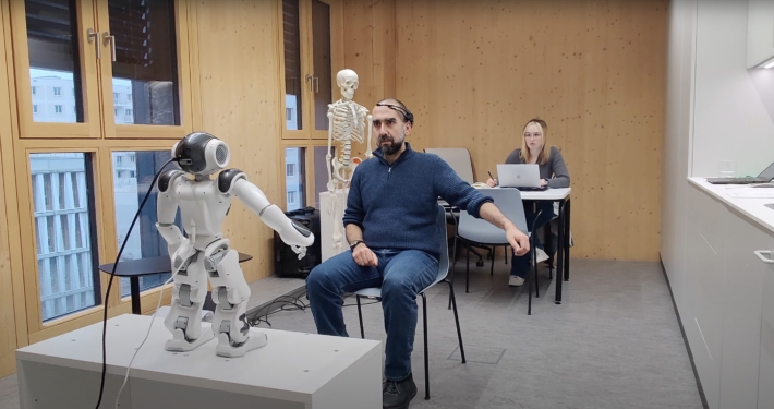 The image shows a humanoid robot interacting with a person wearing VR goggles in an indoor setting.