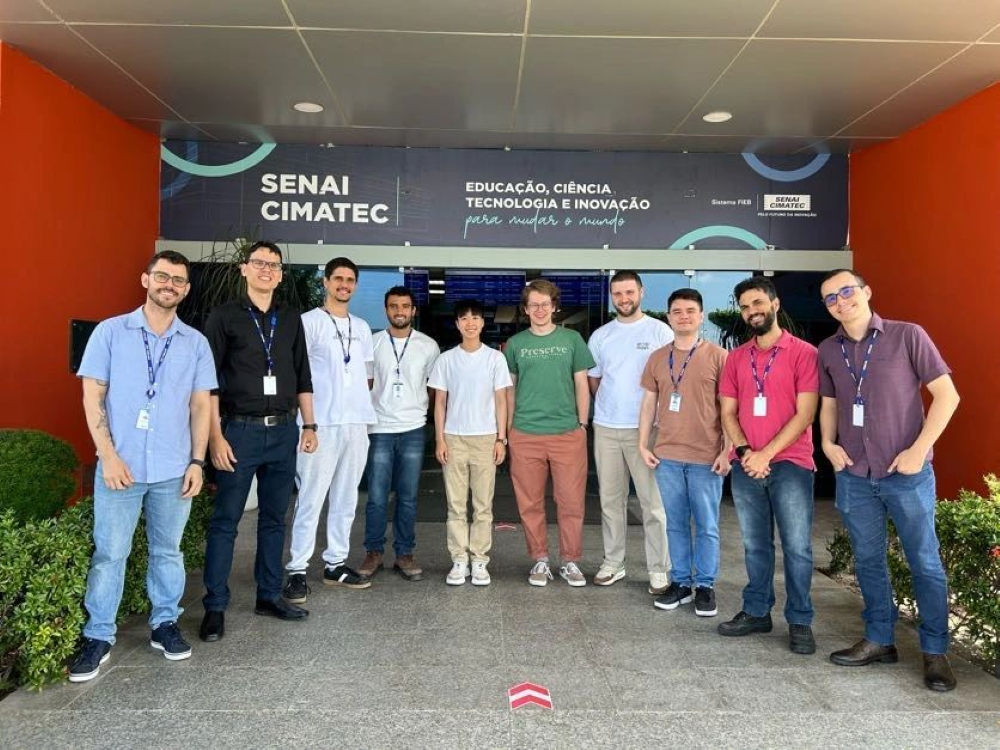 A group photo taken in front of the SENAI CIMATEC building, which has a sign reading "Educação, Ciência, Tecnologia e Inovação para mudar o mundo" (translated as "Education, Science, Technology, and Innovation to change the world").