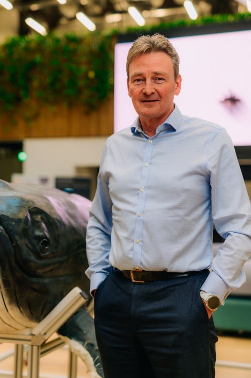 A headshot portrait of a man standing indoors in a modern, well-lit space. He is wearing a light blue button-up shirt and dark trousers, with his hands casually in his pockets. Behind him is a partial view of what appears to be a model of a robotic whale on a stand.