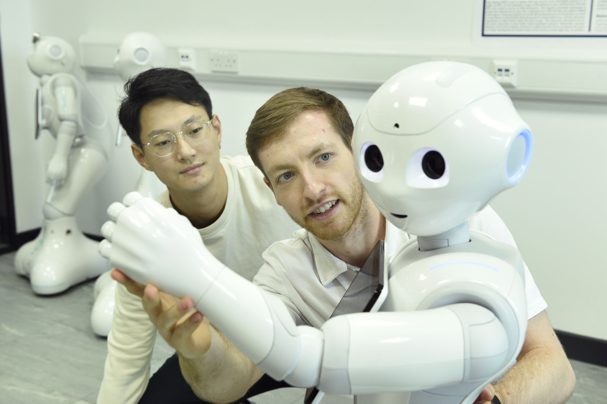 A man moves his arm to mimic a humanoid robot while another man in glasses looks on