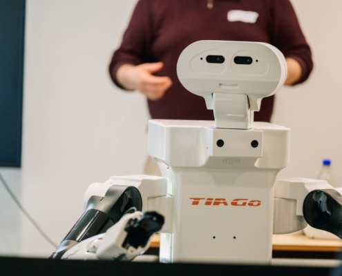 A white humanoid robot labeled "Tiago" is positioned in the foreground, featuring a head with two cameras resembling eyes and articulated robotic arms
