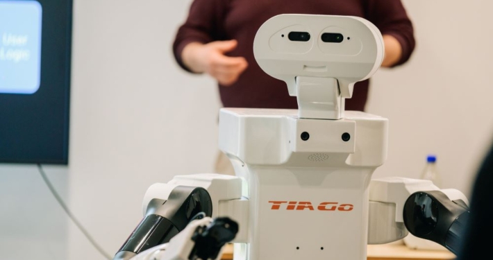 A white humanoid robot labeled "Tiago" is positioned in the foreground, featuring a head with two cameras resembling eyes and articulated robotic arms