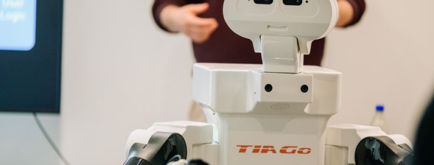 A white humanoid robot labeled "Tiago" is positioned in the foreground, featuring a head with two cameras resembling eyes and articulated robotic arms