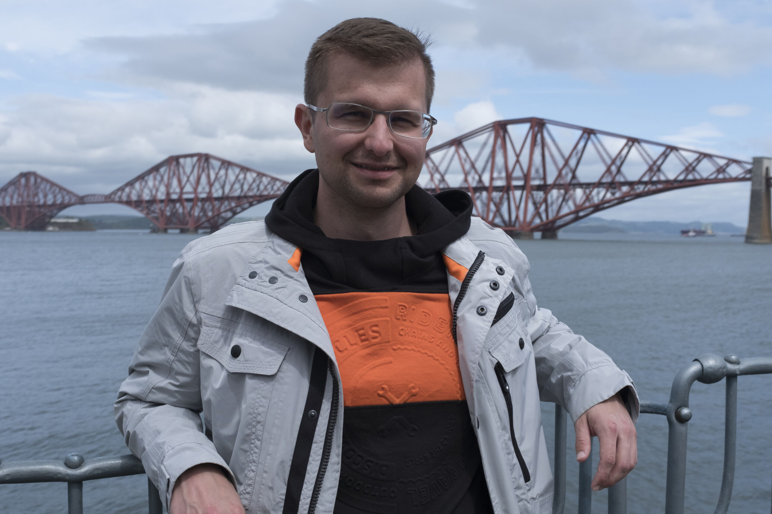 A man with glasses poses in front of the Forth Bridge