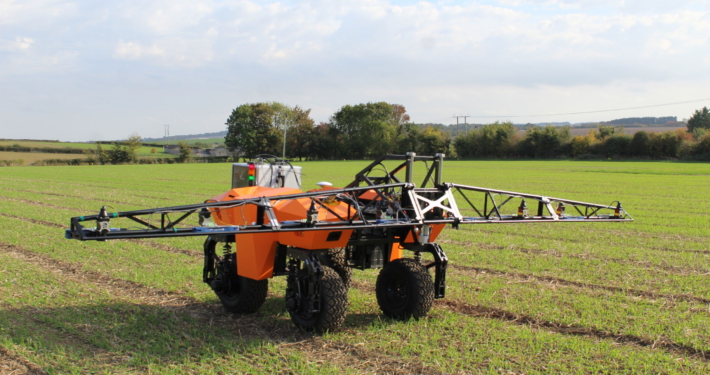 A small orange vehicle on 4 wheels moves across a field. It has a light metal glider on top that stretches out on either side.