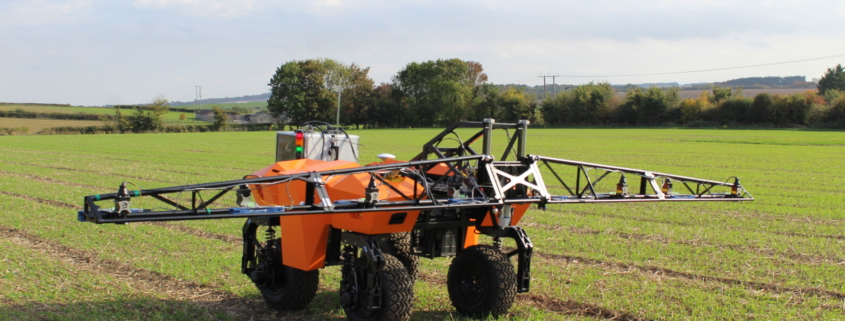 A small orange vehicle on 4 wheels moves across a field. It has a light metal glider on top that stretches out on either side.
